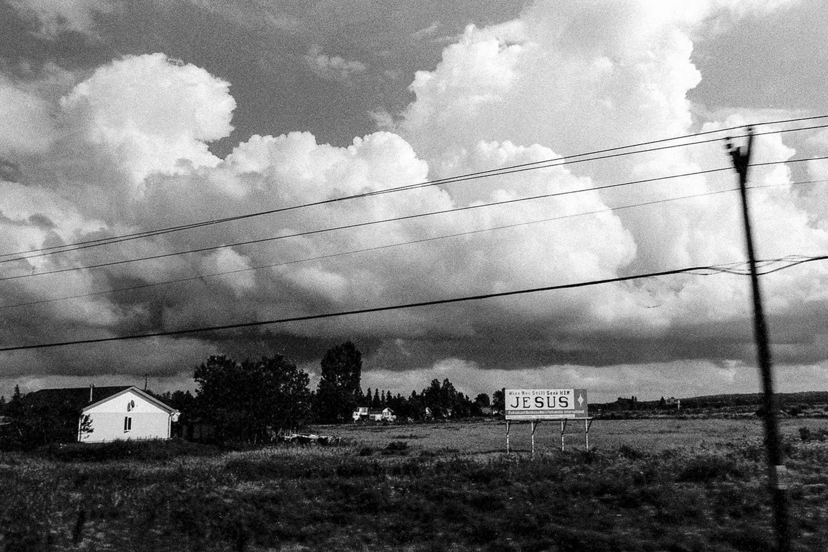 Black and white fine art photography Canada by Ziggy Kańczukowski capturing a rural landscape with a house, trees, and a 'Jesus' sign under a cloudy sky.